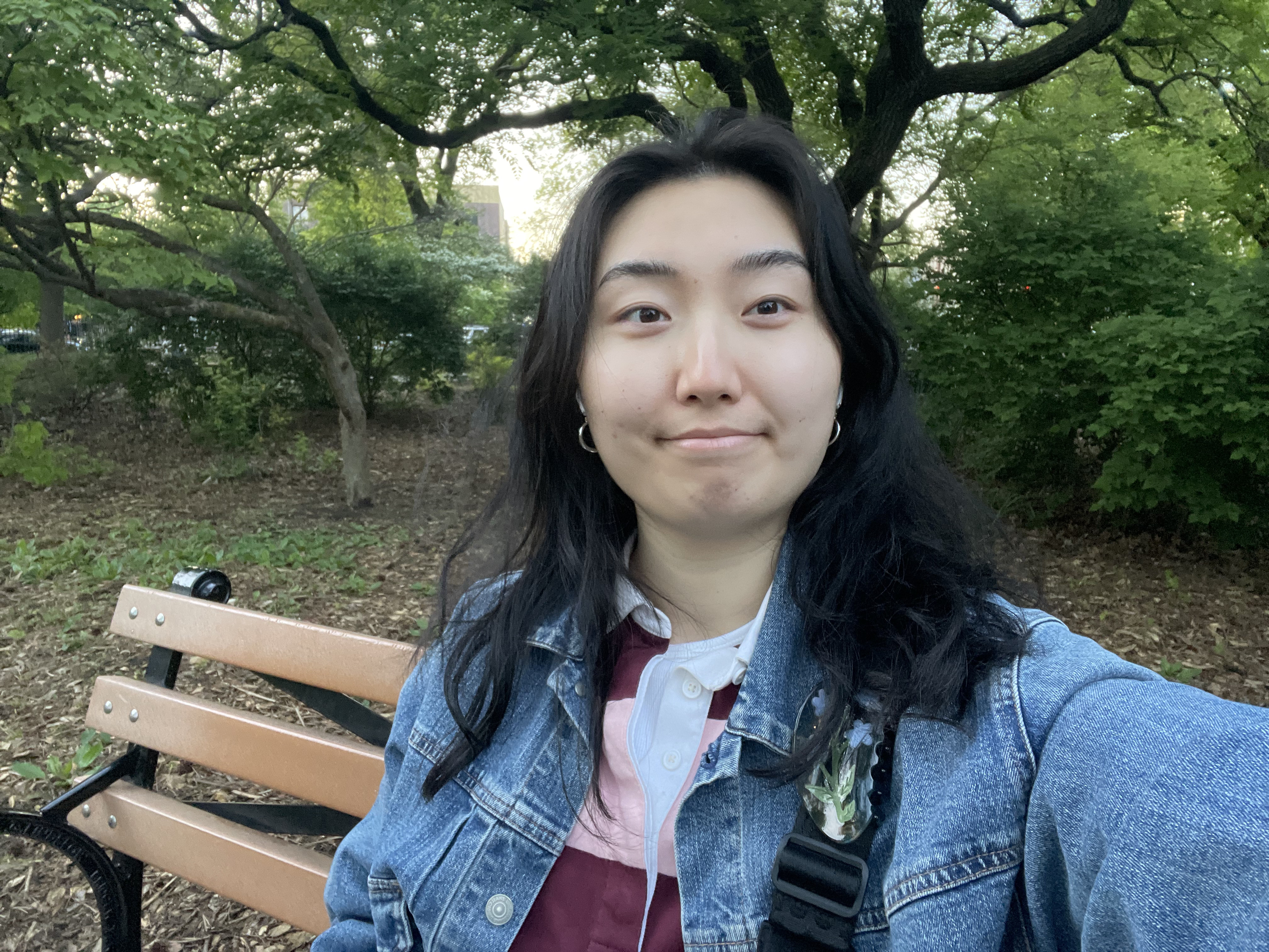 Nicole, an Asian American femme person with wavy black hair, smiles at the camera while sitting on a bench against a background of trees. They are wearing a denim jacket and a striped shirt.