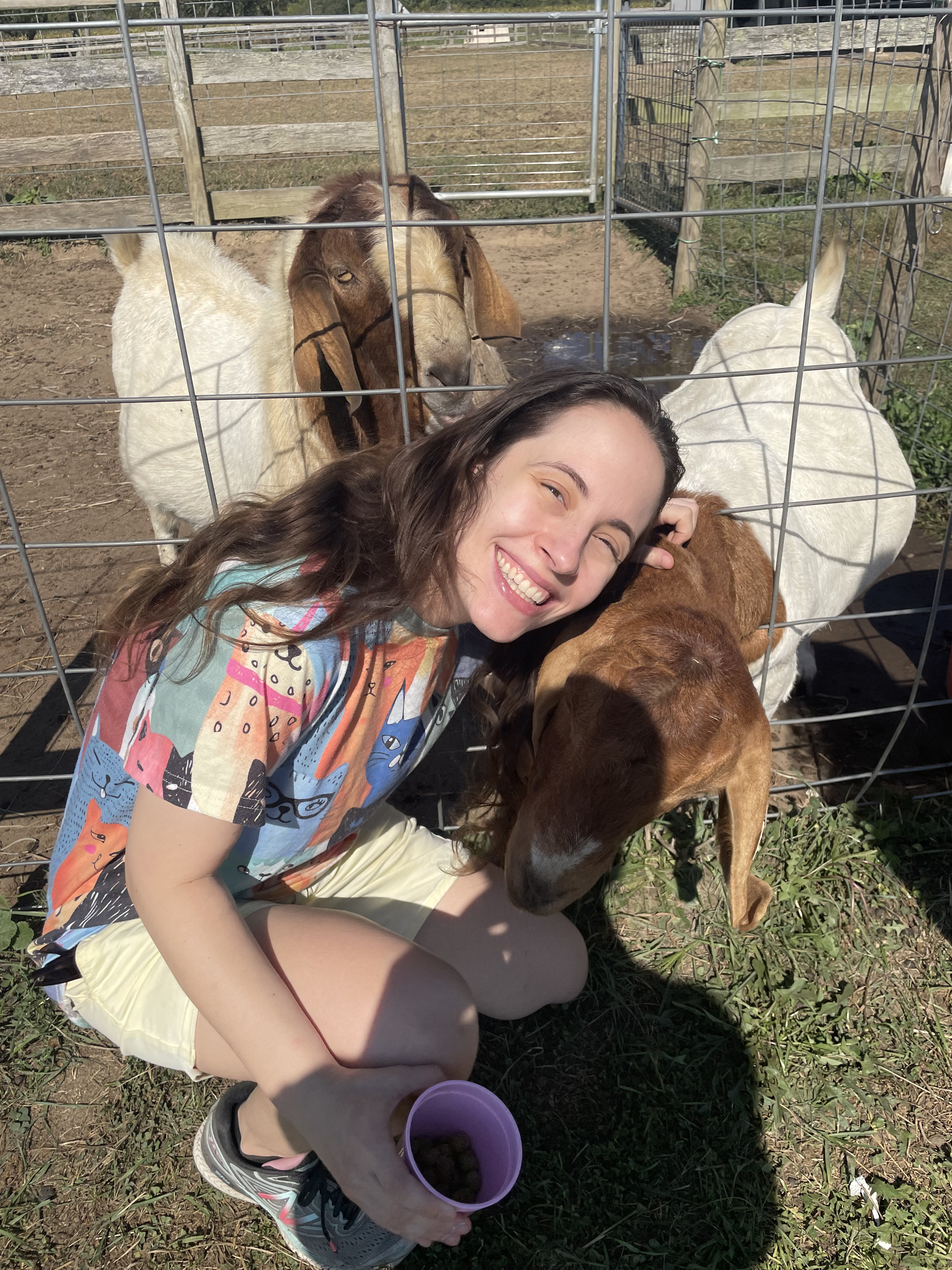 Emily, a white femme person with brown hair, smiles widely at the camera while cuddling a goat. She is wearing a colorful patchwork shirt featuring cats and is crouched down by the goat, bathed in sunlight.